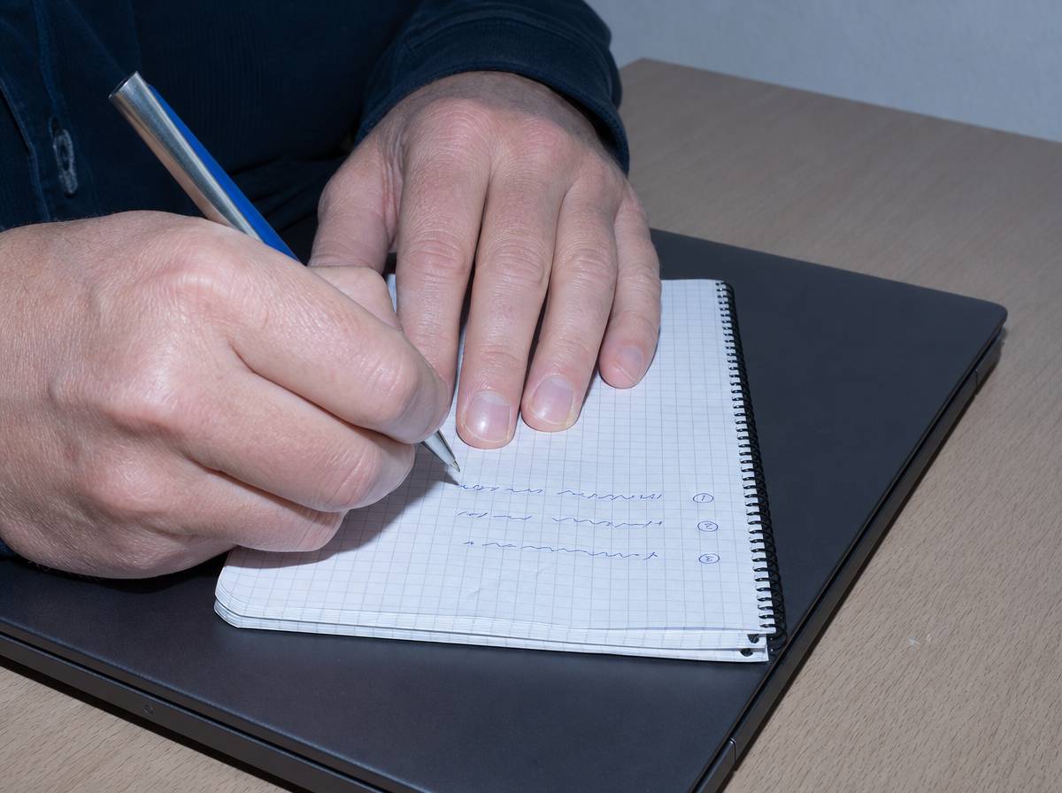 Dłonie zapisujące trzy priorytety dnia w notesie leżącym na zamkniętym laptopie