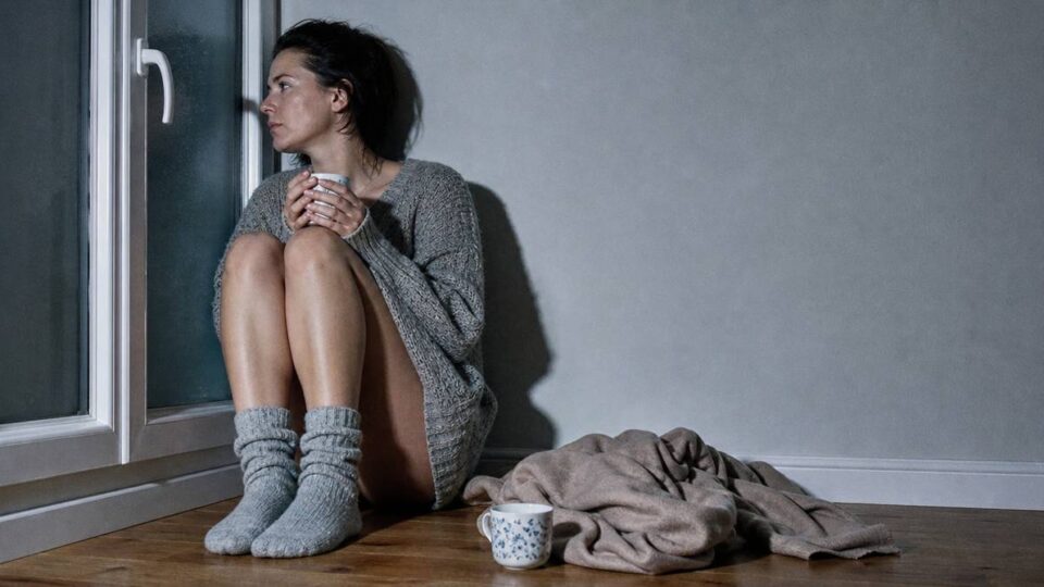 Kobieta w swetrze siedzi przy oknie z kubkiem herbaty, obok nietknięta druga filiżanka — tęsknota za bliską osobą